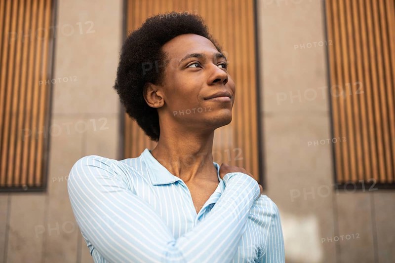 A non-binary individual stands confidently outdoors, adorned in a light blue striped shirt. They gaze upward against a backdrop of modern wood paneling, exuding positivity and self-assuredness