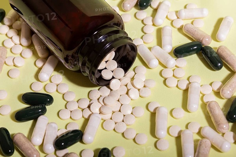 A variety of pills and capsules are scattered from a dark brown bottle onto a light green surface, showcasing a mix of shapes and colors in a healthcare context