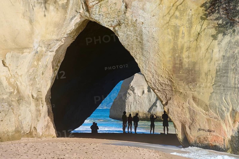 People standing under a large rock arch on the beach overlooking the sea, surrounded by sand and shade, Cathedral Cove, Hahei, Coromandel Peninsula, North Island, New Zealand