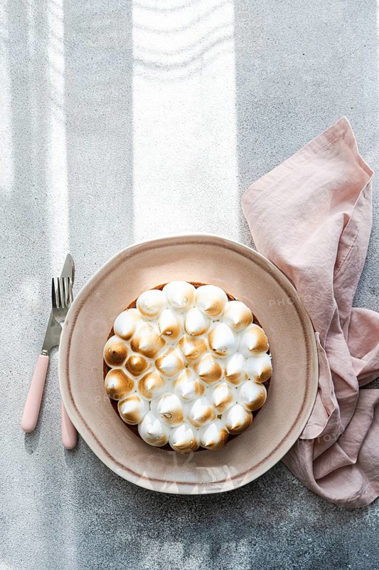A beautifully presented homemade lemon cake on a ceramic plate beside a pink napkin, fork, and knife. Sunlight casts soft shadows over the minimalist setting