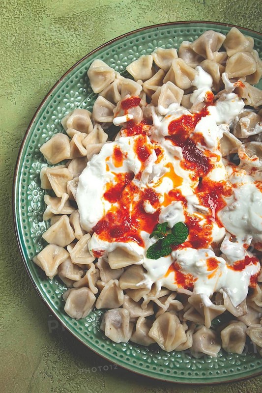The dish features a generous serving of Turkish dumplings topped with velvety yogurt and a drizzle of spicy red sauce, displayed on a beautifully crafted plate with a green accent