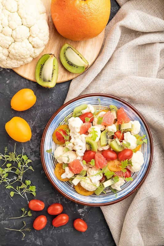 Vegetarian salad of cauliflower cabbage, kiwi, tomatoes, microgreen sprouts on black concrete background and linen textile. Top view, flat lay, close up