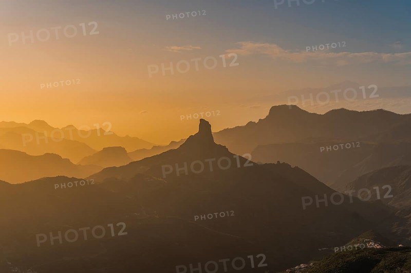 An enchanting sunset view of Roque Nublo standing distinct against a backdrop of layered mountain silhouettes, captured in warm golden and orange hues providing a tranquil and picturesque scene at Gran Canaria