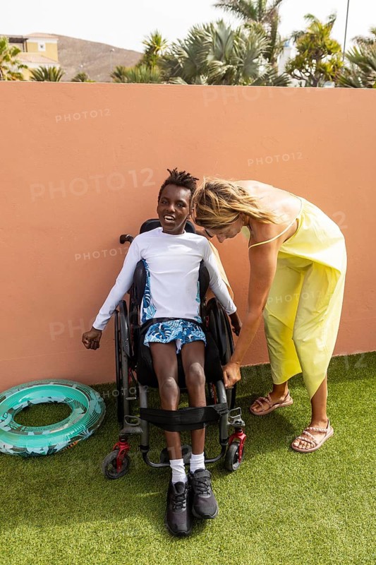 A woman helps a black boy in a wheelchair with cerebral palsy position himself comfortably outdoors. The scene conveys care, support, and inclusive under a sunny sky with lush greenery