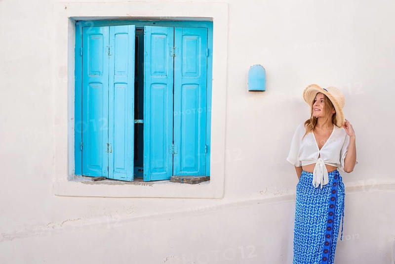 A woman in a straw hat and white blouse stands by a vibrant blue shutter in Trypiti, Milos Island, Greece. The scene captures a serene summer day mood