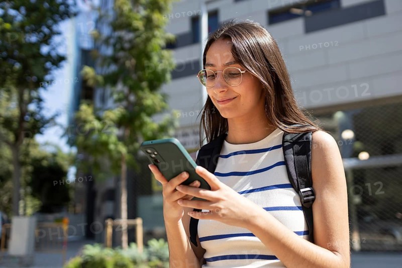 A young woman in casual attire joyfully navigates city life. With a backpack and smartphone, she embodies the blend of technology and urban exploration, smiling under the sun