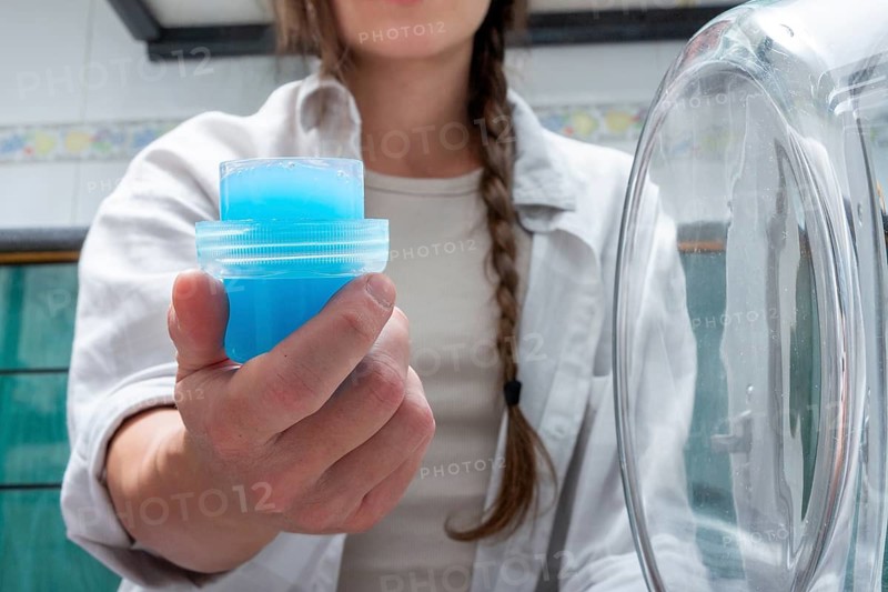 A Gen-Z girl holds a cap full of bright blue liquid detergent next to a washing machine, representing a clean start to a laundry routine in a modern household