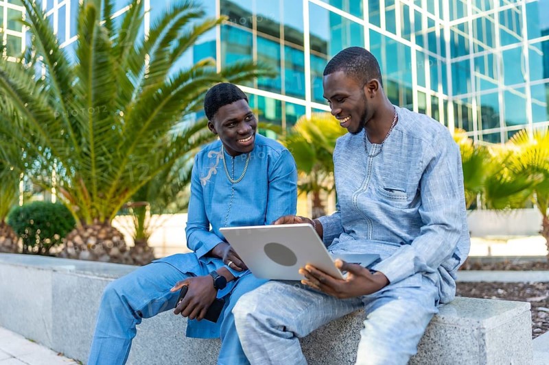 Two senegalese businessmen wearing traditional dashiki attire are using a laptop computer outside a modern office building