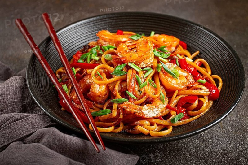 Udon stir-fry shrimp noodles with red bell pepper and green onion, served in a black bowl, with chopsticks, dark background