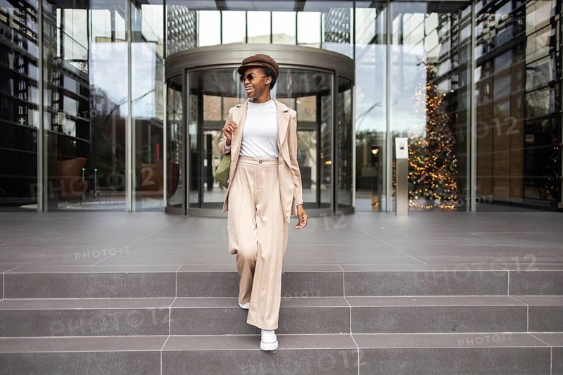 A fashionable black woman with a confident smile, wearing a beige outfit and sunglasses, walking down steps outside a contemporary building Urban lifestyle vibes
