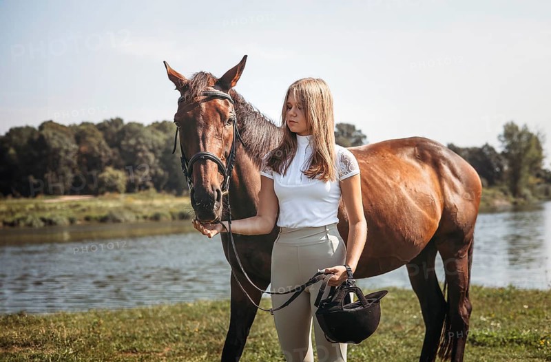 A young girl stands beside a brown horse near a river, holding a riding helmet. The calm outdoor setting highlights equestrian friendship amidst nature tranquility