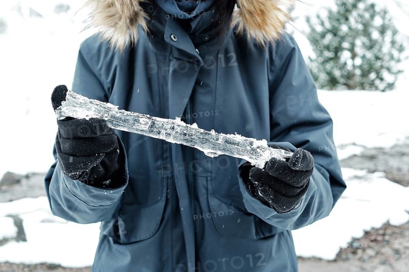 A person wearing a winter coat and black gloves holds a long, textured icicle. The frozen landscape in the background is covered in snow, creating a cold and serene winter atmosphere