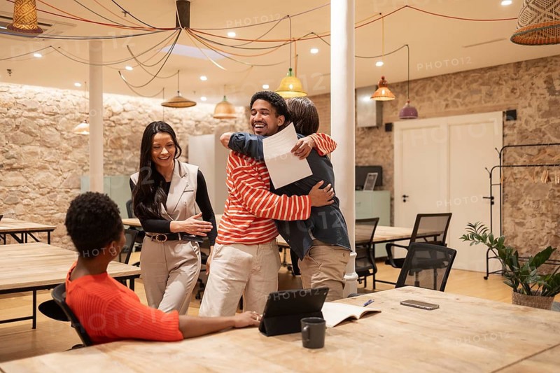 Four colleagues share a joyful moment in a stylish coworking space with exposed stone walls and colorful lighting, emphasizing teamwork and positive work culture