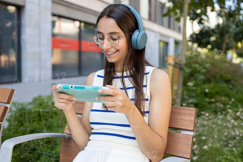 A cheerful woman enjoys her day outdoors in the city, sitting on a bench while wearing headphones and engaging with a handheld device. The setting is lively and urban