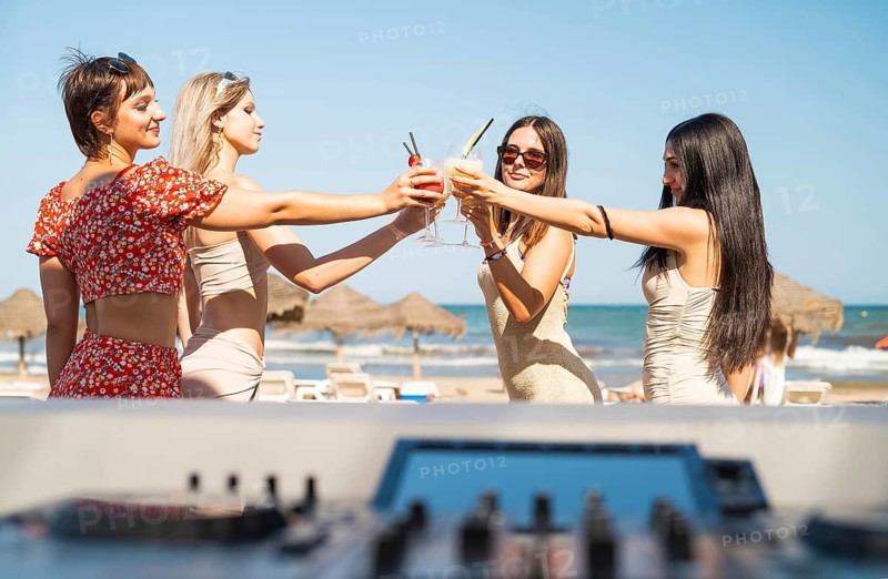 Four women in summer attire raise glasses for a toast at a beach party. The scene captures a festive atmosphere with clear skies, sunlight, and a vibrant coastal backdrop
