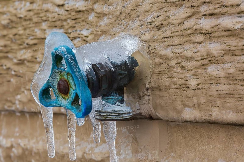 Close-up of outdoor utility brass water faucet with blue handle mounted through Canexel siding on wall of home and covered with ice anf icicles after freezing rain in late winter, Quebec, Canada