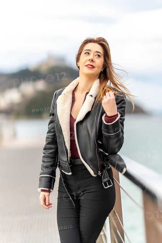 Fashionable woman in a leather jacket gazing at the scenic view from a pier, enjoying the coastal breeze and urban atmosphere
