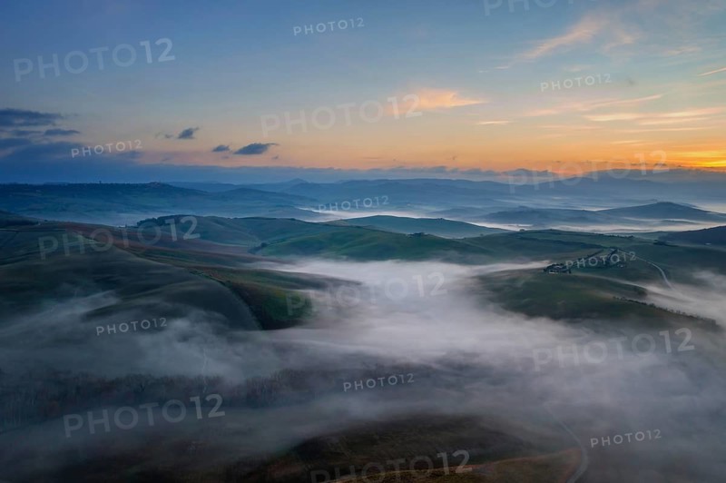 A stunning aerial view of the Val d Orcia in Tuscany, Italy, with rolling hills blanketed in morning mist under a colorful sunrise, creating a tranquil and dreamy landscape