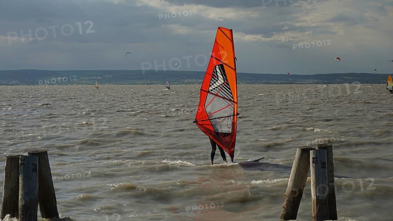 A windsurfer with a red sail stands out against the cloudy sky in the water, Lake Neusiedl, Austria