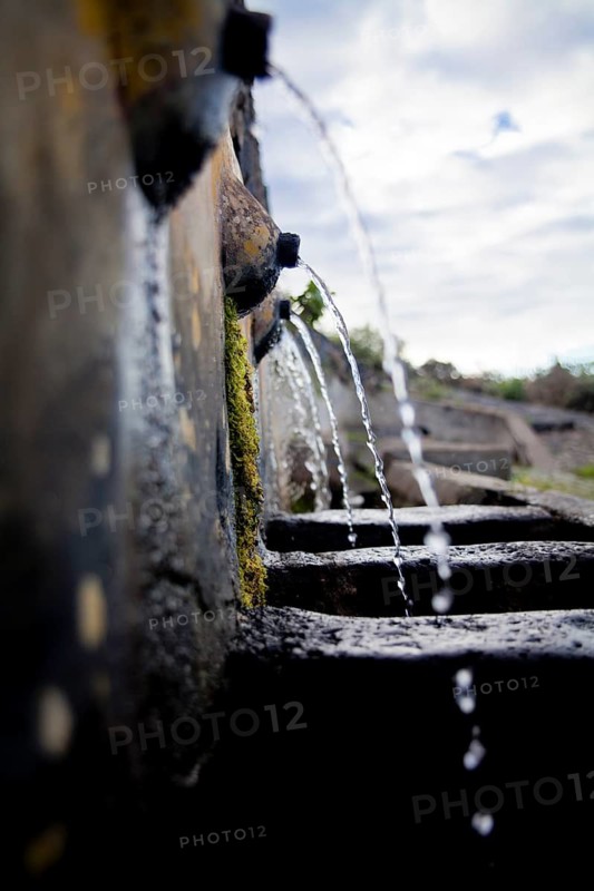 Close-up view captures water flowing through an old, moss-covered stone water channel, symbolizing traditional irrigation methods in El Tanque, Tenerife, with natural lighting enhancing the texture and tranquility of the scene