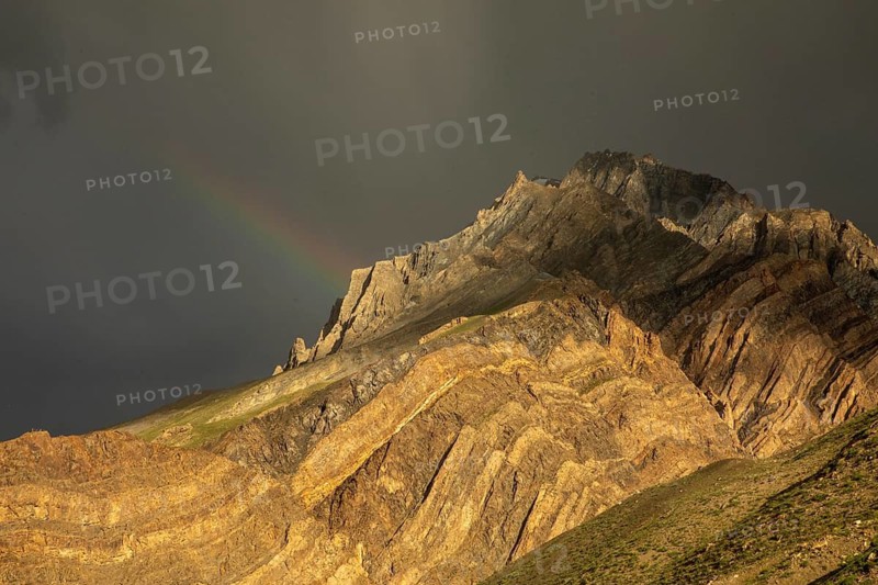 A breathtaking view of a rugged mountain in Spiti Valley, India, adorned with a delicate rainbow against a dramatic sky. The terrain showcases the region untouched beauty