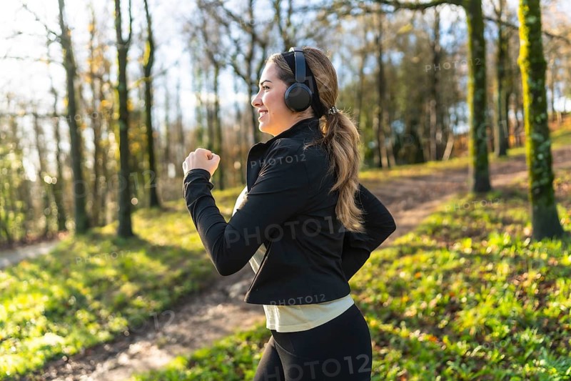 Woman jogging on a leafy park trail wearing headphones and sportswear, smiling as she enjoys a morning run for fitness, fresh air and a healthy, active lifestyle