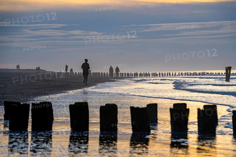 Winter on the North Sea, sunset at the beach, joggers, beach walkers, coastal protection, through wooden posts, paalhoofden, on the beach, breakwater, piles rammed into the sandy soil in double rows slow down the waves and ocean currents and thus slow down the flow rate of seawater, so less sand is washed away from the standing position, near Domburg, Walcheren peninsula in the Dutch province of Zeeland