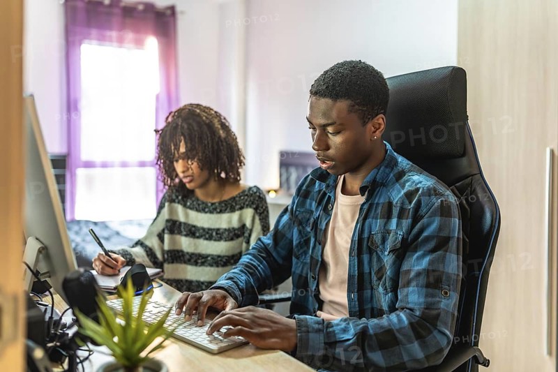 Two focused young students are studying together at home, using a desktop computer and taking notes