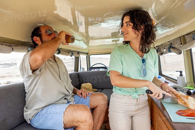 A blind man and a woman in a motorhome, enjoying a relaxing moment. He is sipping a drink while she prepares a meal, symbolizing adventure and companionship