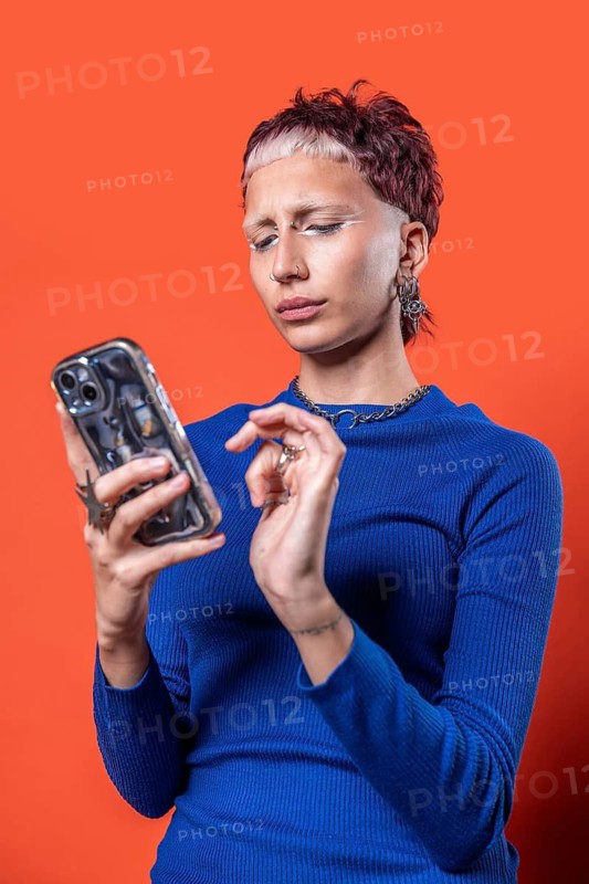 A stylish woman in a blue top looks at her smartphone against a bold orange backdrop. The scene captures modern fashion with a focus on her edgy hairstyle and accessories