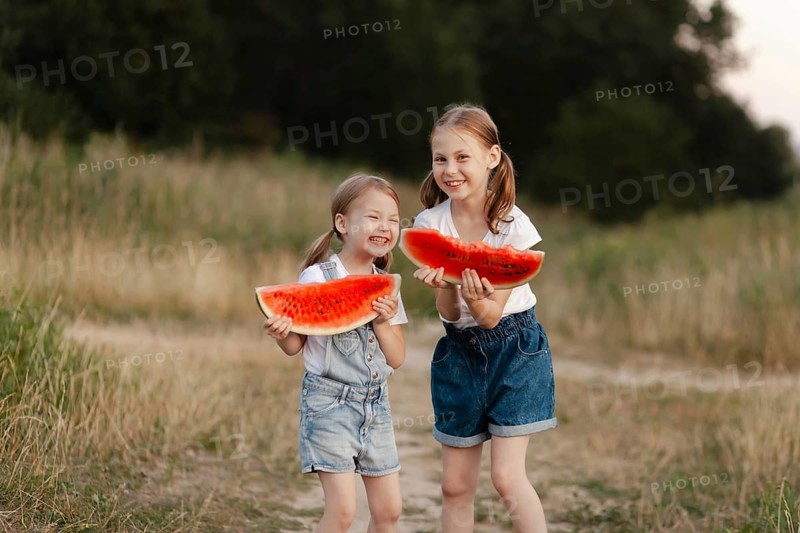 Two smiling children hold juicy watermelon slices in a sunlit field during summer. The scene captures the joy of a warm day and the refreshing taste of watermelon outdoors