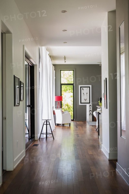 Main hallway with Qumaru wood floorboards leading to living room with white leather armchair, red floor lamp inside a luxurious modern cube style house, Quebec, Canada