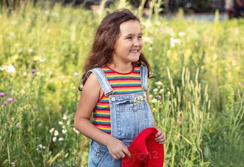 A cheerful little girl stands smiling in a sunlit meadow, holding a chamomile flower. Her playful expression captures the essence of joyful childhood moments in nature