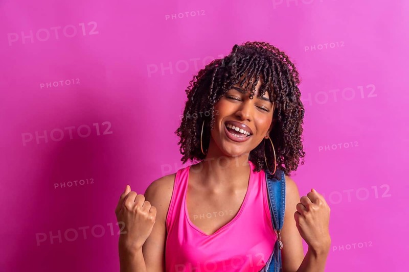 Young woman with curly hair, wearing a pink top and denim overalls, joyfully celebrating against a vibrant pink backdrop