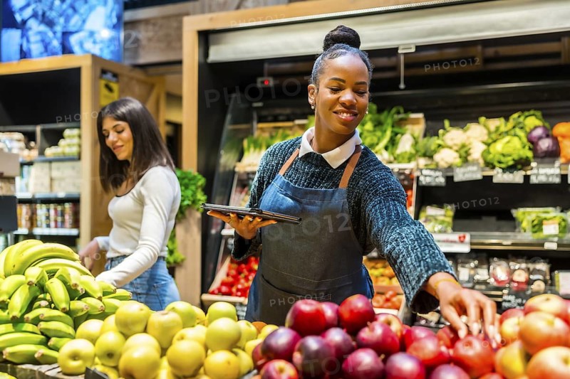 Smiling saleswoman arranging fresh apples and using digital tablet while customer choosing bananas in supermarket