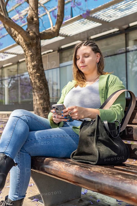 Young woman with braided hair, green jacket, and black bag using her smartphone on a sunny day. She sits on a wooden bench surrounded by blooming jacaranda trees in an urban setting