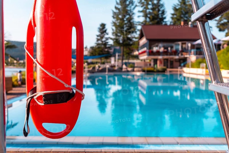 A red life preserver is hanging on a metal pole next to a pool. The scene is calm and peaceful, with the water reflecting the sky and the surrounding trees