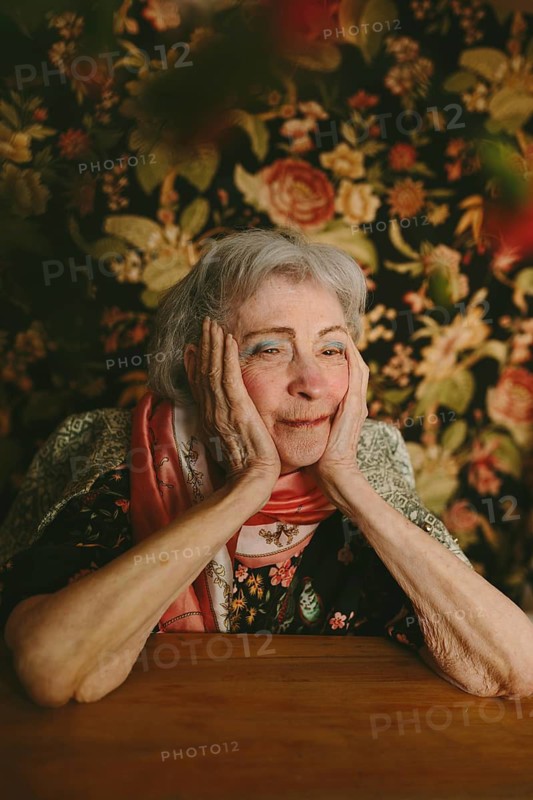 An elderly woman sits thoughtfully at a table, her hands gently resting on her face, surrounded by a rich floral backdrop, evoking nostalgia and warmth in a studio setting