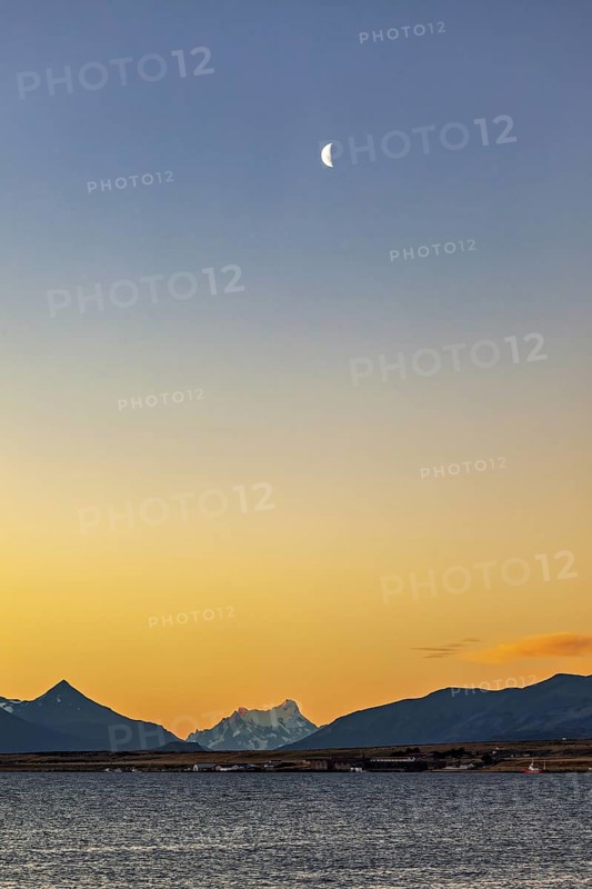 Half moon in the sky over a calm lake surrounded by mountains at dusk, sunset at the lake near Puerto Natales in Patagonia Chile