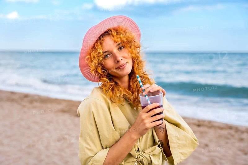 Curly haired woman wearing a pink straw hat enjoying a refreshing drink on the beach during a summer day