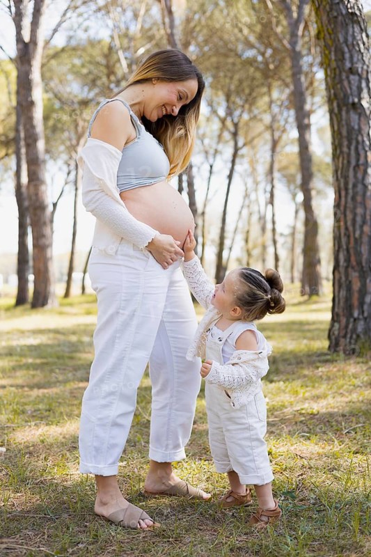 A pregnant woman stands in a serene forest scene with her young daughter, who gently touches her mother's belly. The joyful and loving moment captures family and expectation