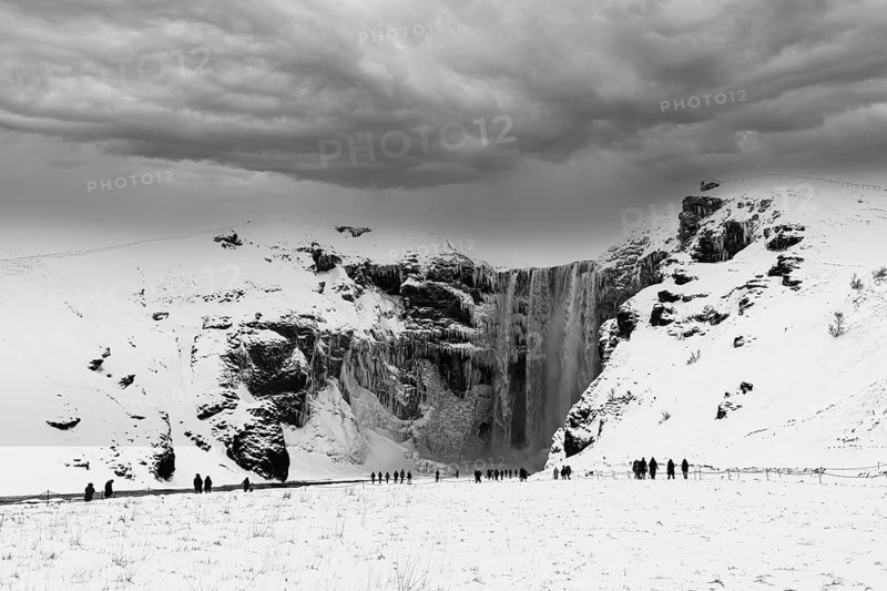 Tourists at Skogafoss waterfall, snowy and icy rock face, dark clouds above, black and white photo, Sudurland, Iceland