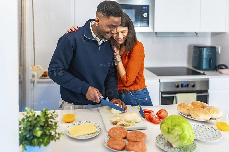 Young multiethnic couple hugging and slicing onions, preparing burgers for dinner in a modern white kitchen
