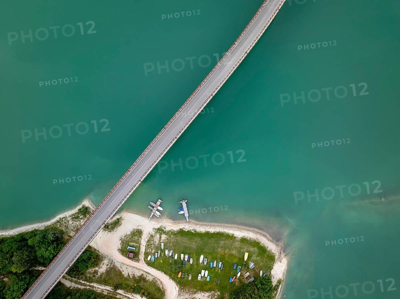 Top-down drone image of Castreccioni Lake showing a bridge crossing turquoise water and a small beach area with docked boats, creating a calm, organized and natural summer landscape