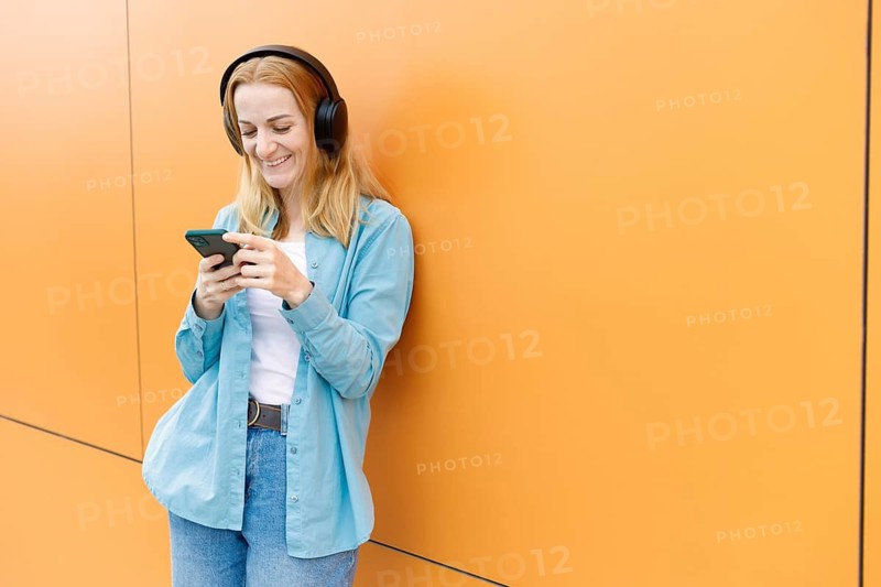 Smiling woman leans against a vibrant orange wall in Warsaw, listening to music on wireless headphones and using her smartphone Casual attire enhances her modern vibe