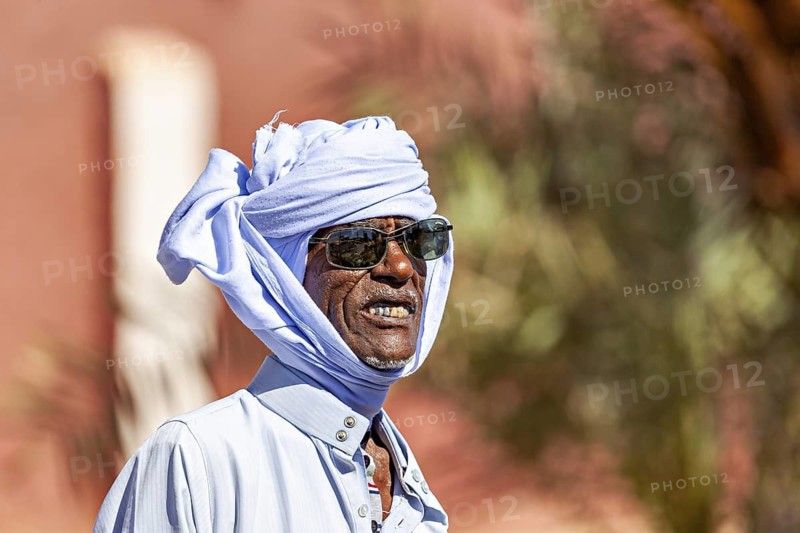 A man with sunglasses and turban smiles in the sun, dressed in a light blue shirt, man with sunglasses and turban in Algeria