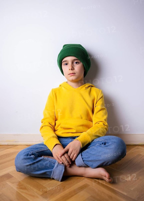 A young kid sits cross-legged on a wooden floor, wearing a vibrant yellow hoodie and green beanie. The child looks pensive while leaning against a white wall