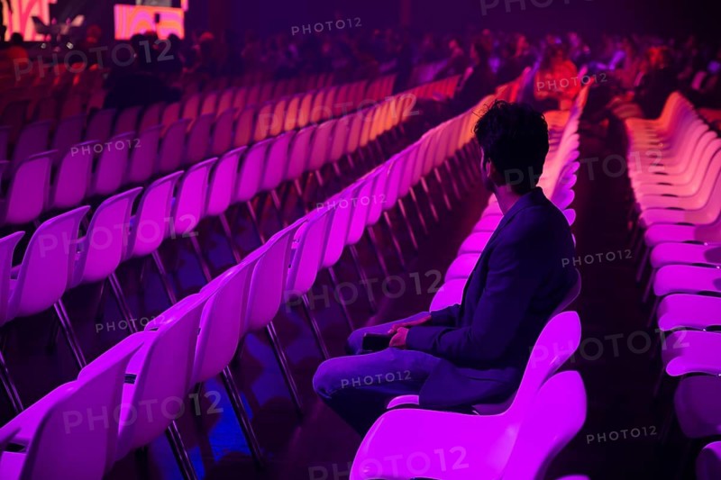 A indian man in a vast auditorium under dim purple lighting at a conference of AI in Amsterdam. Rows of empty chairs surround him, creating a sense of anticipation