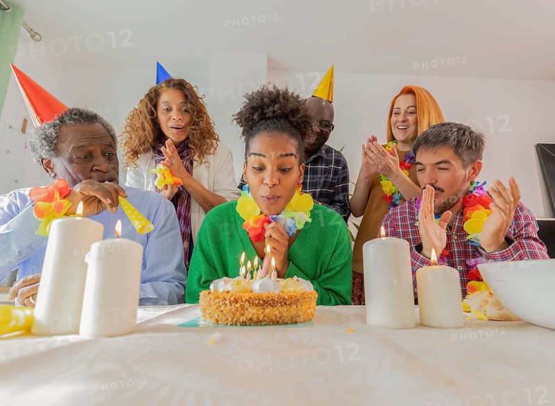 Group of friends celebrating a birthday with a cake and lit candles. Everyone is wearing party hats and leis, enjoying the festive atmosphere and joyful occasion