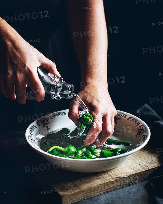 Hands gently wash fresh green peppers in a decorative bowl, under a stream of water from a bottle, capturing a moment of vegetable preparation in a rustic kitchen setting